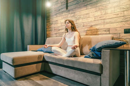 Young calm woman in lotus pose practice yoga or relaxing meditation on sofa at home in evening.の写真素材