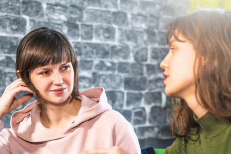 Two girls talks, gossiping or chatting. One of young woman looks at her friend with smile and...