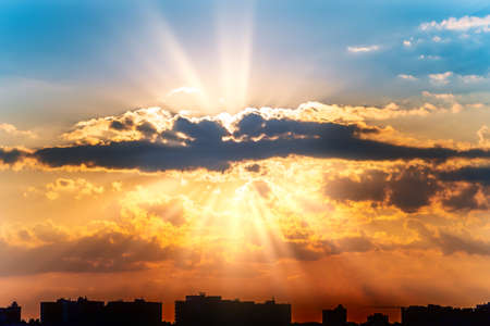 Colorful dramatic summer sunset with rays of sun from clouds over city buildings. Beautiful sunshine in heaven.の写真素材