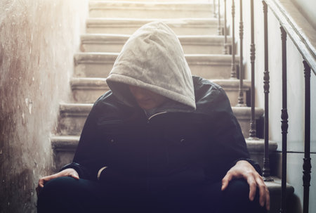 Lonely man in hood with his head down sits on stairs in dark. Stress and depression mental problems concept.の写真素材