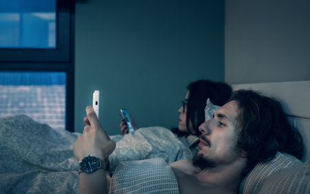 Married couple lying in bed together but each of partners use smartphone. Man and woman looks at...