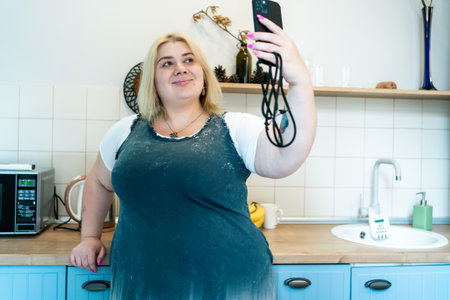 Positive Plus Size Blonde young woman making video call or selfie by mobile phone on kitchen at home.の写真素材