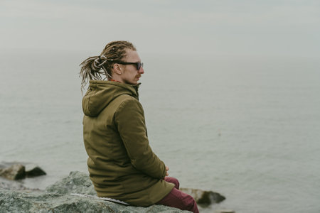 Lonely man sits by sea shore in cloudy, gloomy weather. Sadness, depression, and mental problems concept, copy spaceの写真素材