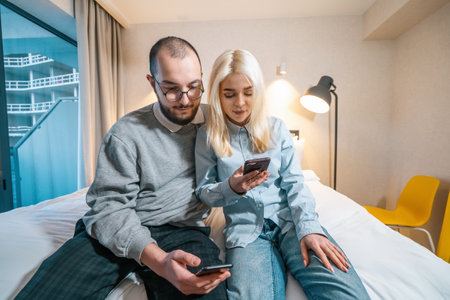 Young couple each looking at their phone while sitting on bed at home spending time together wife and husband.の写真素材