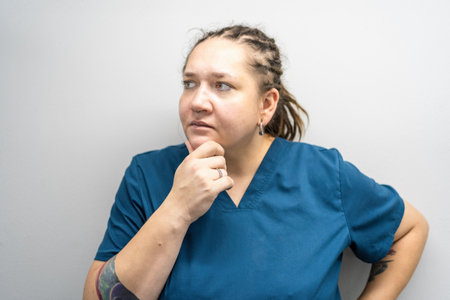 Plus size woman in nurse uniform looks thoughtfully to side, scratching her chin. Making difficult decisions concept.の写真素材