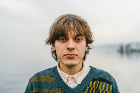 Young male with contemplative gaze, wearing sweater, against blurred lakeside backdrop.の写真素材