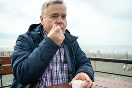 Senior man savoring ice cream while sitting at beachfront cafe, contemplating and enjoying view.の写真素材