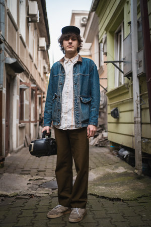 Stylish young man with vintage cassette player in old city alley. Modern retro fashion in hip culture. Vertical imageの写真素材