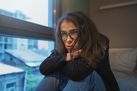 Thoughtful young woman with glasses sitting by window, looking lost in contemplation, with...