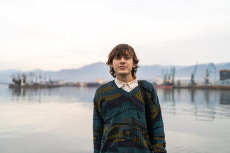 Young man in vintage inspired attire stands before tranquil harbor backdrop, fusing classic style...