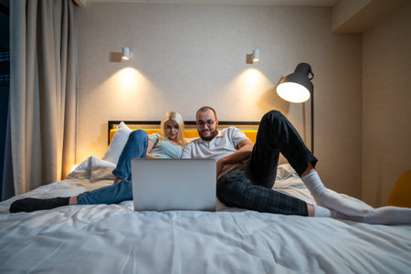 Couple lies on bed, sharing moment of connection over laptop screen, in cozy bedroom setting.の写真素材