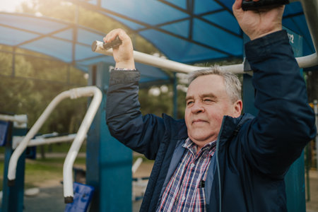 Senior man exercising outdoor fitness equipment in park. Elderly gentleman using pull-up bars under...