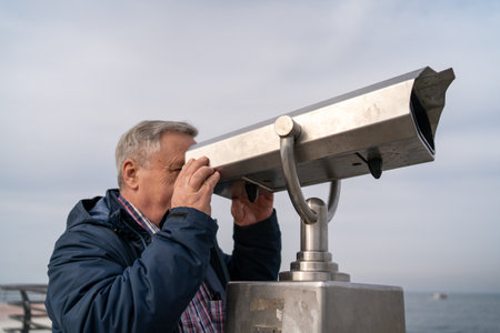 Senior man using public telescope at seaside. Elderly gentleman observing distant view through...
