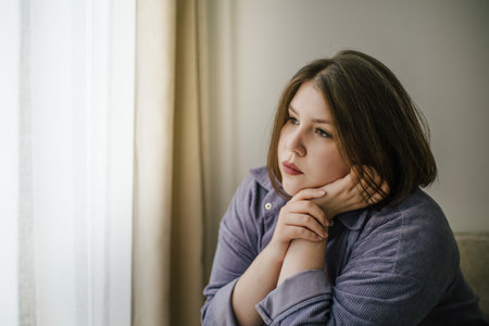 Sad plus-size woman with pensive expression looking out window with sheer curtains.の写真素材