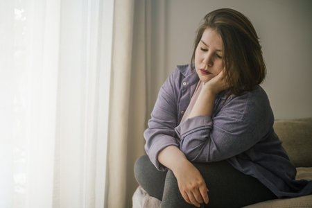 Plus size woman sitting alone by window, wearing casual clothes.の写真素材