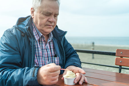 Older man savoring cup of ice cream while sitting on bench near seaside on cloudy day.