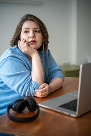 Plus-size young woman rests her head on her hand, looking thoughtfully into distance. She seated at desk with laptop and headphones.の写真素材
