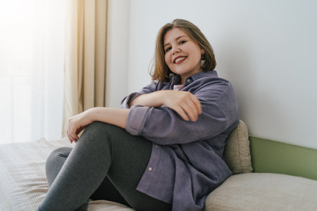 Smiling plus-size woman sitting on bed, looking at camera with relaxed and confident expression. Cozy home environment, self-acceptance and positivity. Casual and comfortable lifestyle.の写真素材