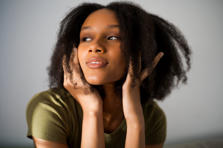 Young African American woman with curly hair, resting chin in hands, looking up thoughtfully. She exudes sense of contemplation and tranquility against light background.の写真素材