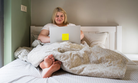 Positive plus size woman using laptop in bed, enjoying a relaxed and cozy atmosphere. Promoting remote work, body positivity, and comfortable home lifestyle.の写真素材