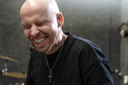Man laughing with eyes closed in a music studio. Close-up portrait with grey background and drum equipment in the scene.の写真素材