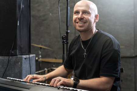 Bald male musician playing keyboard in studio, smiling with joy, wearing black clothing, surrounded by recording equipment.の写真素材