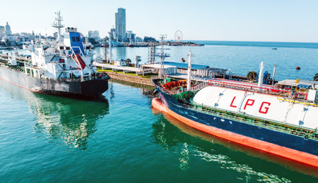 LPG tanker and cargo ship near industrial port with urban skyline in background, captured from elevated angle. Reflection on water highlights maritime trade and energy logistics.の写真素材