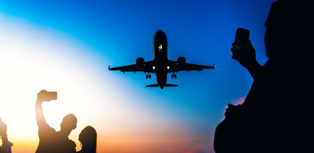 Airplane silhouette in sunset sky with unrecognizable silhouettes capturing moments on smartphones, symbolizing travel spirit and modern connectivity.の写真素材