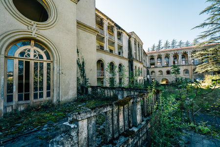 Exterior of abandoned neoclassical building with arched windows, intricate facade details, overgrown plants, and weathered stone elements, blending historical architecture with nature reclaiming.の写真素材