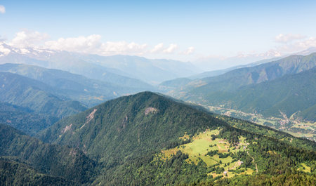 Aerial view of a vast mountain landscape covered in dense green forests and rolling hills. Small rural villages are scattered across the valley, blending with nature under a clear blue sky.の写真素材