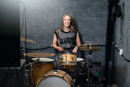 Smiling female drummer sitting behind drum set in music studio. Positive vibe, passion for music, and creativity in rehearsal environment.の写真素材