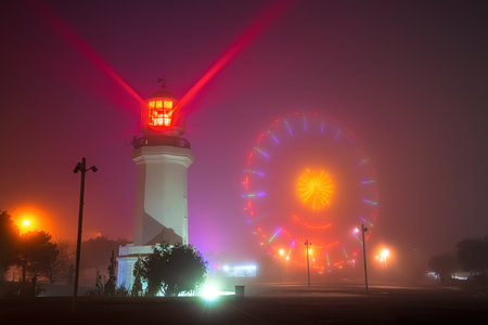 Mystical foggy night in Batumi with glowing lighthouse casting red beam beside vibrant Ferris wheel illuminated in colorful lights. Perfect for travel, mystery, or coastal themes.の写真素材