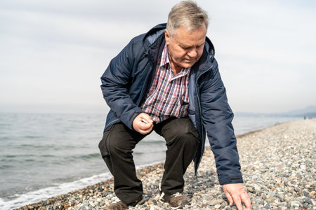 Older male bending down to collect rocks on a pebble beach, engaging with nature. Represents...
