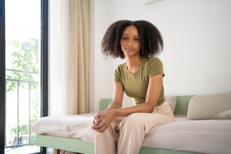 Dark-skinned woman with natural curly hair sitting on bed in cozy room. Lifestyle, mindfulness, comfort and authenticity concepts.の写真素材