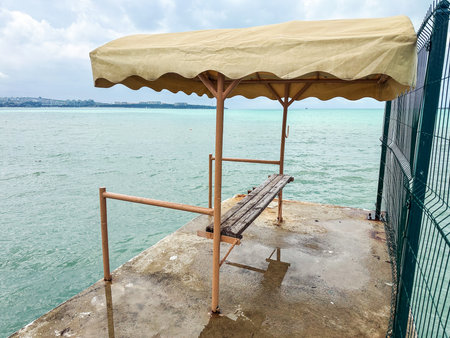 Old wooden bench with yellow canopy on waterfront pier under cloudy sky, evoking solitude, reflection, and travel inspirationの写真素材