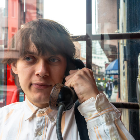 Young man using vintage payphone in city booth, expressing nostalgia, retro communication, emotional...