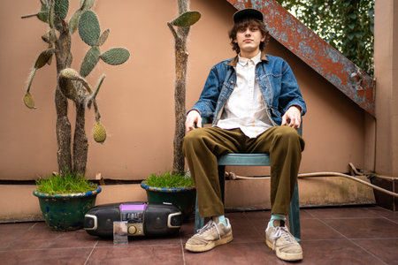 Young man in retro outfit with cassette player and cactus pots sitting in urban backyard. Vintage...