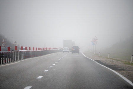 Vehicles moving along highway in thick fog with low visibility and traffic signs, illustrating dangerous driving conditions and road safety in poor weather.の写真素材
