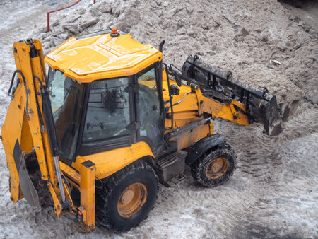 Yellow municipal tractor removing dirty snow in apartment courtyard during winter, concept of public...
