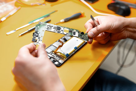 Smartphone repair in a service center. Hands of a professional master repairman repairing disassembled into pieces cell phone. Internal parts, chips, motherboards, wiring, fixing process.の写真素材