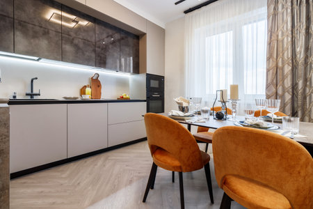 Modern kitchen combined with the dining room in dark and light colors, with orange chairs and glass cabinets for dishes.の写真素材