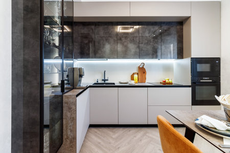 Modern kitchen combined with the dining room in dark and light colors, with orange chairs and glass cabinets for dishes.の写真素材