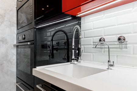 Kitchen faucet is black, near the wall with glossy white bricks and glossy black and red cabinets.の写真素材