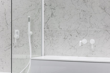 Modern bathroom with white faucets and shower, walls lined with white marble.の写真素材