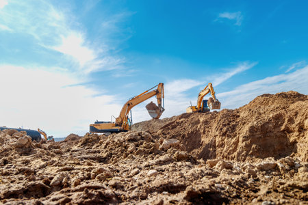 Two Excavators are digging soil in the construction site on sky background,with white fluffy cloud.の写真素材