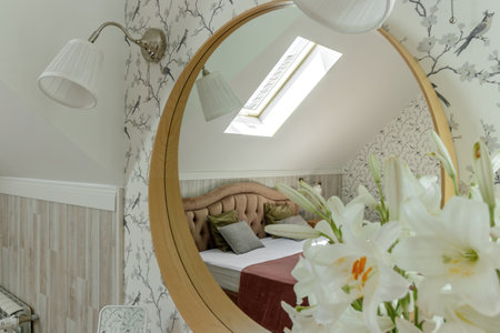 The reflection in the round mirror reveals a charming attic bedroom adorned with floral wallpaper, soft pillows, and fresh flowers, making it a serene and inviting spaceの写真素材