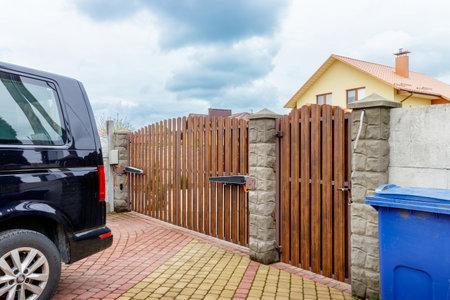 A wooden gate with stone pillars stands at the entrance of a driveway, with a black car parked nearby, surrounded by a neatly paved path.の写真素材