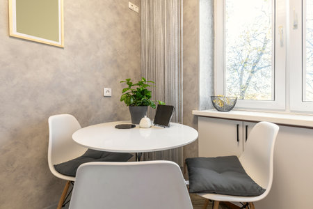 A modern kitchen nook featuring white minimalist furniture, a small round table, and a green plant...