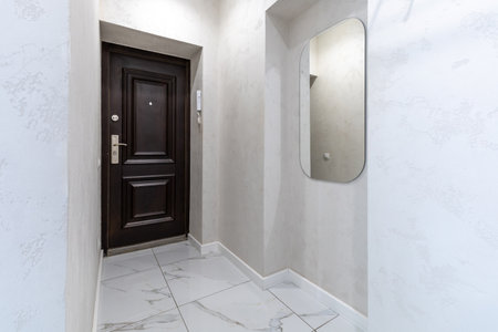 A modern minimalist entryway featuring a dark wooden door, an arched mirror, light walls, and marble flooring for a sleek and elegant look.の写真素材