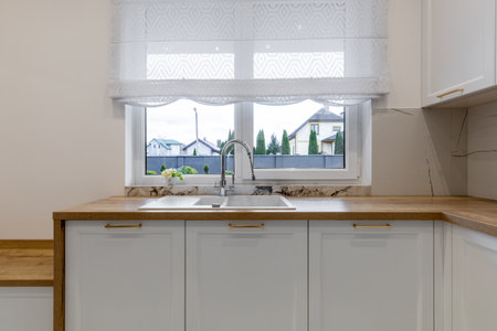 A stylish kitchen sink with a chrome faucet set on a wooden countertop, placed under a window with elegant white blinds, offering a scenic outdoor view.の写真素材